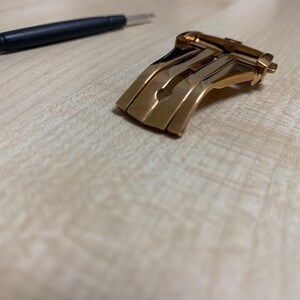 Peut inclure: Un fermoir de montre doré et un outil à manche noir sur une surface en bois clair. Le fermoir a un design incurvé avec plusieurs barres parallèles et un mécanisme à charnière. L'outil est fin et métallique.