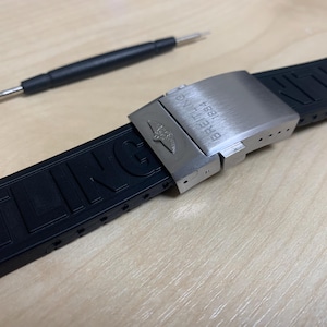 May include: A black rubber watch strap with a stainless steel clasp, featuring the Breitling logo and text. A small watch tool is positioned above the strap. The strap is designed for a watch.