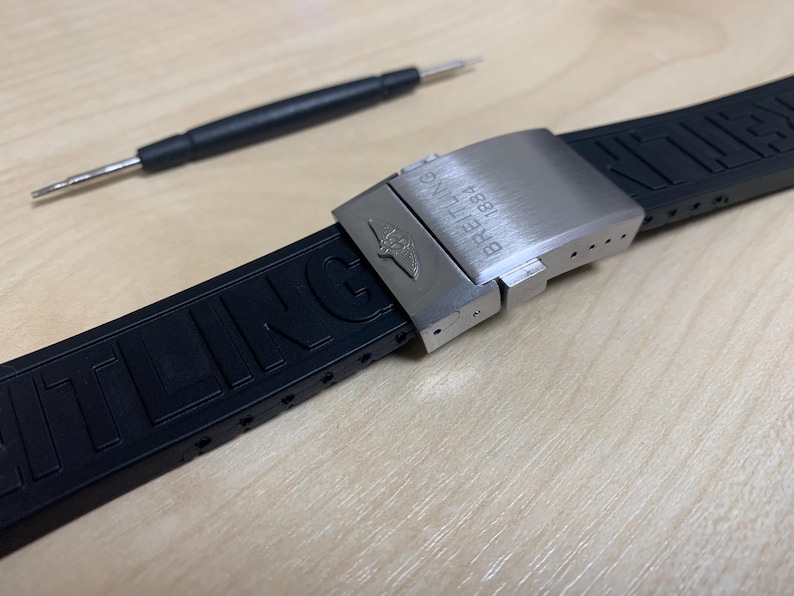 May include: A black rubber watch strap with a stainless steel clasp, featuring the Breitling logo and text. A small watch tool is positioned above the strap. The strap is designed for a watch.