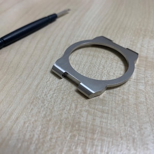 May include: A silver-colored watch bezel and a watch tool are displayed on a light-colored wooden surface. The bezel is circular with rectangular extensions. The tool has a black handle and a silver tip.