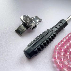 May include: A silver watch clasp with the Longines logo, a black watch repair tool, and a strand of pink beads. The watch clasp is made of shiny metal. The repair tool has a black handle and a silver tip.