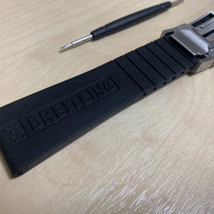 May include: Black watch strap with the brand name "BREITLING" embossed on the strap. The strap is made of a flexible material and has a stainless steel clasp. A small screwdriver is also in the image.