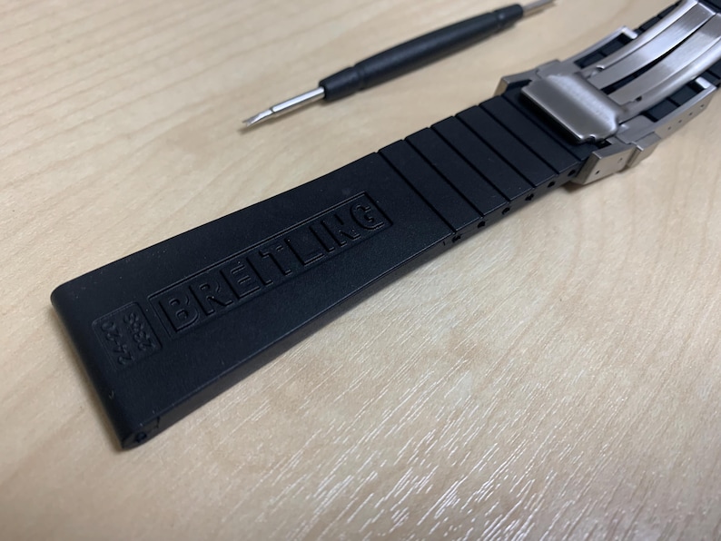 May include: Black watch strap with the brand name "BREITLING" embossed on the strap. The strap is made of a flexible material and has a stainless steel clasp. A small screwdriver is also in the image.