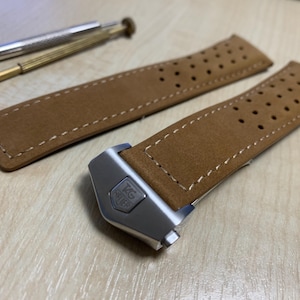 May include: A tan leather watch strap with white stitching and a silver clasp. The strap has small perforations and is accompanied by two small screwdrivers, likely for watch repair or adjustment.