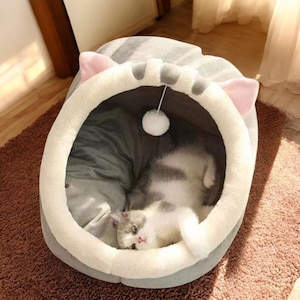 Könnte beinhalten: Eine Katze liegt in einem Katzenbett in Form eines Katzenkopfes. Das Bett hat eine grau-weiß gestreifte Außenseite, eine weiß-graue Innenseite und rosa Ohren. Ein weißer Pompon-Spielzeug hängt von oben.