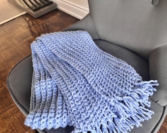 Puede incluir: Una manta tejida a mano de color azul claro con flecos. La manta está colocada sobre un sillón gris tapizado. La textura tejida es visible y los flecos cuelgan sueltos en el borde de la manta.