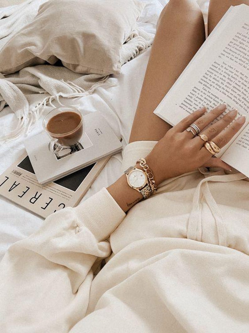 Op de afbeelding: Een ontspannen sc&egrave;ne met een kopje koffie, boeken en een polshorloge. Het horloge heeft een gouden en zilveren band en een ronde wijzerplaat. De boeken hebben de titels "PARIS" en "CEREAL" op de covers. De sc&egrave;ne speelt zich af op een bed met een cr&egrave;mekleurige deken en kussen.