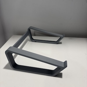 May include: A gray laptop stand with a minimalist design. The stand is made of a solid material and features a rectangular base with angled supports. It is designed to elevate a laptop for improved ergonomics.