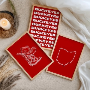 May include: Three rectangular framed prints with light wood frames. One print features the word BUCKEYES repeated in white text on a red background. Another print shows a white line drawing of a cowboy boot and hat on red. The third shows a white outline of the state of Ohio on red.