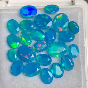 Pode incluir: Uma coleção de opalas ovais em vários tamanhos e tons de azul e verde-azulado. Algumas opalas exibem flashes de vermelho, laranja e verde. As pedras preciosas estão dispostas sobre uma superfície branca.