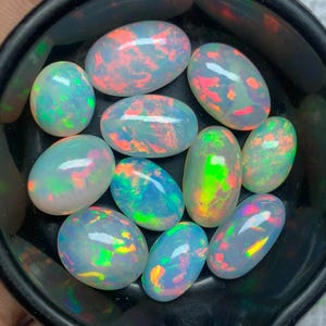 Puede incluir: Una colección de ópalos ovalados en un cuenco negro. Los ópalos muestran una gama de colores, incluyendo blanco, azul, verde, naranja y rojo, con destellos iridiscentes. Las piedras preciosas son lisas y pulidas.