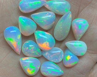 May include: A collection of opalescent gemstones in various teardrop and oval shapes. The stones display a play of color, with flashes of green, orange, and blue within a milky white base. The gemstones are held in a hand.