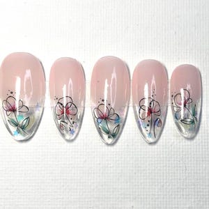 May include: Set of five artificial nails with a light pink base and a floral design. Each nail features a hand-drawn flower with black outlines, red and blue accents, and a clear, glossy finish. The nails are almond-shaped.