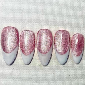 May include: Five artificial nails with a pink glitter top and a white base. The nails are oval-shaped and vary in size, with the largest nail on the left and the smallest on the right. The glitter reflects light.