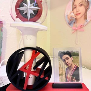 May include: A white and red light stick with a star design, a black and red stand with the text "YOU MAKE STRAY KIDS STAY", and a photo card of a man in a clear case. A fan with a photo of a person is in the background.