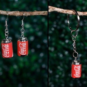 Może przedstawiać: Kolczyki i brelok w kształcie miniaturowych puszek Coca-Coli. Kolczyki i brelok przedstawiają małe, szczegółowe repliki kultowych czerwono-białych puszek Coca-Coli. Kolczyki wiszą na srebrnych haczykach, a brelok ma srebrny pierścień.