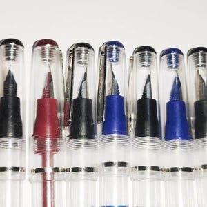 Könnte beinhalten: Eine Sammlung klarer Füllfederhalter mit verschiedenfarbigen Tintenbehältern und Kappen. Die Stifte haben schwarze, blaue und rote Tinte, mit transparenten Schäften und silbernen Akzenten. Die Kappen sind transparent, sodass man die Federn sehen kann.