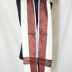 May include: A white scarf with a brown paisley pattern along the edges and down the center. The scarf has a fringed edge and is displayed on a black mannequin. The scarf is made of a soft material.