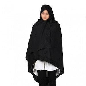 May include: A black shawl with intricate embroidered detailing. The shawl is draped over a person wearing a black head covering and black pants, with a white shirt visible underneath. The edges of the shawl feature a decorative border.