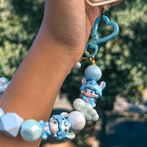 May include: A light blue beaded phone charm bracelet with cartoon character accents. The bracelet features a variety of light blue beads, including round, iridescent, and geometric shapes. The charm includes a blue heart-shaped clasp and a cartoon character.