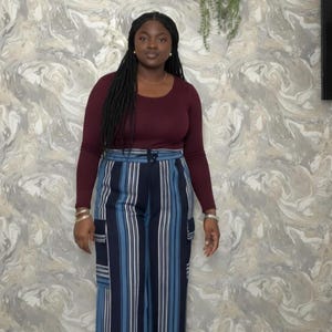 May include: A woman wearing a burgundy long-sleeve top and wide-leg pants with vertical stripes in shades of blue and white. She is also wearing white sneakers. The background features a marbled wall.