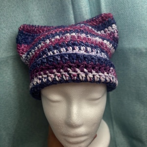 May include: A handmade crochet cat ear hat in shades of blue, purple, and white. The hat is displayed on a white mannequin head. The hat features a striped pattern and is made of soft yarn.