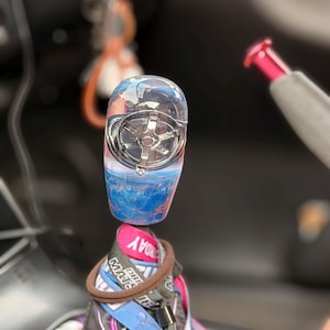 Peut inclure: Un pommeau de levier de vitesse cylindrique en résine transparente avec un emblème de roue à cinq branches argenté incrusté à l'intérieur. La base du pommeau présente un motif tourbillonnant bleu et blanc. Il est fixé sur un levier de vitesse.
