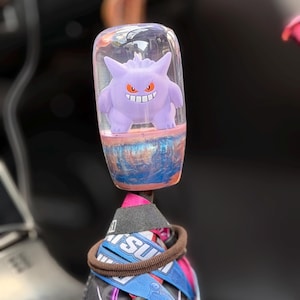 Könnte beinhalten: Ein zylindrischer Schaltknauf mit einem oberen Teil aus klarem Harz, der eine violette Gengar-Figur mit roten Augen und einem breiten Grinsen enthält. Der untere Teil des Knaufs hat eine wirbelnde, blau-kupferfarbene Metallic-Oberfläche.
