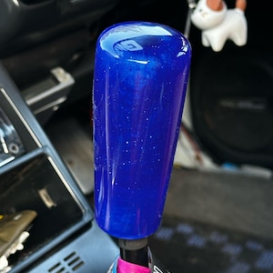 Op de afbeelding: Een blauwe, cilindrische versnellingspookknop met een glanzende afwerking en een gespikkelde uitstraling. De basis van de knop is bevestigd aan een zwarte as met een roze en blauw label. Een kleine honden sleutelhanger hangt op de achtergrond.