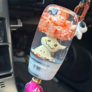 Op de afbeelding: Een doorzichtige hars schakelpookknop met een wit en geel cartoonfiguur en een rood en oranje wolkenachtig ontwerp aan de binnenkant. De knop heeft een wit en grijze basis en een goudkleurige metalen fitting.