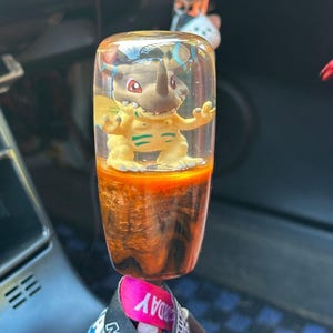 May include: A custom shift knob with a cartoon character in clear resin. The knob has an orange and brown marbled base. It is attached to a lanyard with the text "DRIFT MATSU".