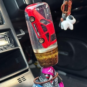Puede incluir: Un pomo de palanca de cambios de coche personalizado con un coche de juguete rojo encapsulado en resina transparente. El pomo tiene una base dorada y está unido a una palanca de cambios con varios llaveros y accesorios.