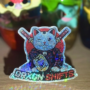 Puede incluir: Una pegatina holográfica con un gato guiñando un ojo con un atuendo de samurái, con espadas cruzadas detrás. La pegatina tiene el texto "DRXGN SHIFTS" en la parte inferior y un logotipo en la barriga del gato. El gato es azul con un atuendo rojo y negro.