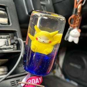 Op de afbeelding: Een helder, rechthoekig auto-accessoire met een gele Pikachu-figuur in een blauwe vloeistof met glitter. Het accessoire heeft een rood en wit lint met het woord "RDAY". Een bruine sleutelhanger met een witte kattenhanger is zichtbaar.