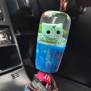 Op de afbeelding: Een aangepaste auto versnellingspookknop met een Bulbasaur figuur ingekapseld in heldere hars. De knop heeft een blauw en groen kleurenschema, met een wervelend effect in het blauwe gedeelte. De basis van de knop is bevestigd aan een verzameling kleurrijke polsbandjes.