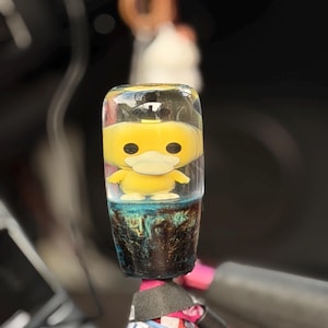 Peut inclure: Un pommeau de levier de vitesse cylindrique en résine transparente contenant un personnage de dessin animé jaune au bec large. La base du pommeau présente un motif marbré bleu et noir sombre. Une sangle en tissu bleu avec du texte blanc est fixée.