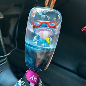 Puede incluir: Pomo de palanca de cambios de coche transparente teñido de azul con una figura de Stitch con gafas de sol rojas y sacando la lengua. El pomo tiene una base veteada azul y negra y está sujeto a un cordón.