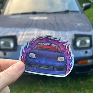 May include: A sticker depicting a blue sports car with the text "DRXGN SHIFTS GARAGE" in a flame design. The sticker is held in front of a blue car.