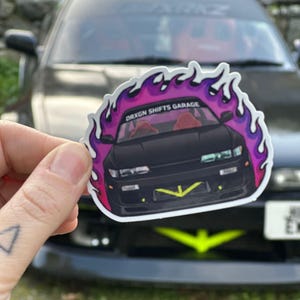 May include: A sticker featuring a black car with a neon green front bumper and red interior, surrounded by a purple and pink flame design. The text "DRXGN SHIFTS GARAGE" is printed on the sticker.