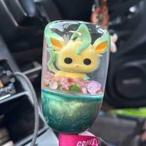 Könnte beinhalten: Individueller Autoschaltknauf mit einer gelb-grünen Cartoonfigur in einem transparenten Harzgehäuse. Der Sockel ist türkisgrün, mit rosa und lila Blumendekorationen. Ein einzigartiges Autozubehör.