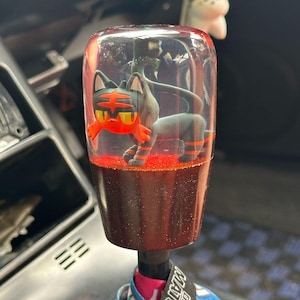 Puede incluir: Pomo de palanca de cambios de coche cilíndrico transparente con una figura de gato de dibujos animados en el interior. El gato tiene marcas naranjas y negras. La base del pomo es marrón oscuro con purpurina roja.