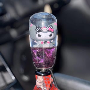 Puede incluir: Un pomo de palanca de cambios cilíndrico transparente con un personaje de dibujos animados con un lazo negro y rosa. El pomo tiene un líquido morado y arremolinado en la base. El pomo de la palanca de cambios está sujeto a una tela estampada en rojo y blanco.