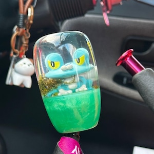 May include: A clear, cylindrical gear stick knob featuring a blue frog figurine. The base is bright green, and the frog has yellow eyes. A pink and black lanyard hangs below the knob.