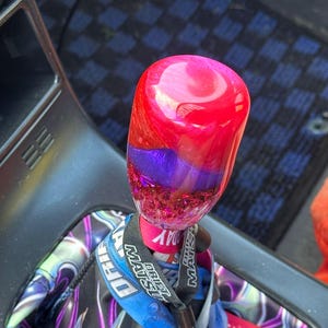 May include: A custom shift knob with a vibrant, layered design. The top is a translucent red, transitioning to purple and a glittery base. The knob is attached to a shift lever with a blue and white lanyard.