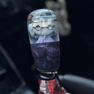 Puede incluir: Un pomo de cambio personalizado con una figura en su interior, con barba blanca, gafas y sombrero, encapsulado en resina transparente. La parte inferior tiene un diseño de remolino púrpura y azul. Adjunto a una tela estampada en rojo y blanco.