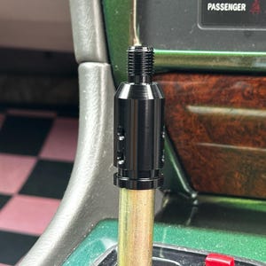 May include: A black cylindrical metal gear shift adapter with a threaded top section and three small side holes, mounted onto a metallic rod. The adapter features a smooth finish and a stepped design, positioned vertically within a vehicle interior.
