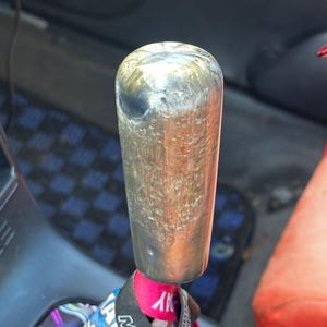 May include: A silver gear stick knob with a textured finish. The knob is cylindrical with a rounded top. A pink and blue fabric tag is attached to the base of the knob.