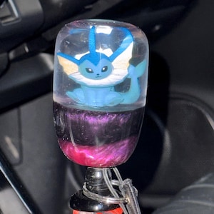 May include: A custom shift knob featuring a blue Vaporeon character encased in clear resin. The bottom portion of the knob has a purple and pink galaxy design. A black base and a red and white patterned fabric are visible.