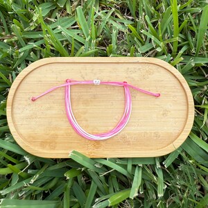 May include: A pink and white friendship bracelet with adjustable ties, resting on a small, oval wooden tray. The bracelet features several strands of pink and white cord, knotted together. The tray is made of light-colored wood.