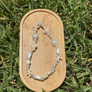 May include: A beaded phone charm featuring white pearls, starfish, seashells, and turtle charms. The charm is displayed on a small, light brown wooden tray, set against a backdrop of green grass.